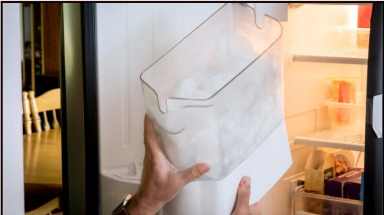 Hands removing full ice bin from refrigerator freezer compartment during ice maker maintenance with visible shelves and food items inside.