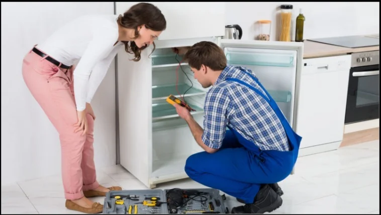 Technician using multimeter to diagnose refrigerator issue while homeowner watches in modern kitchen with tools spread on floor nearby.