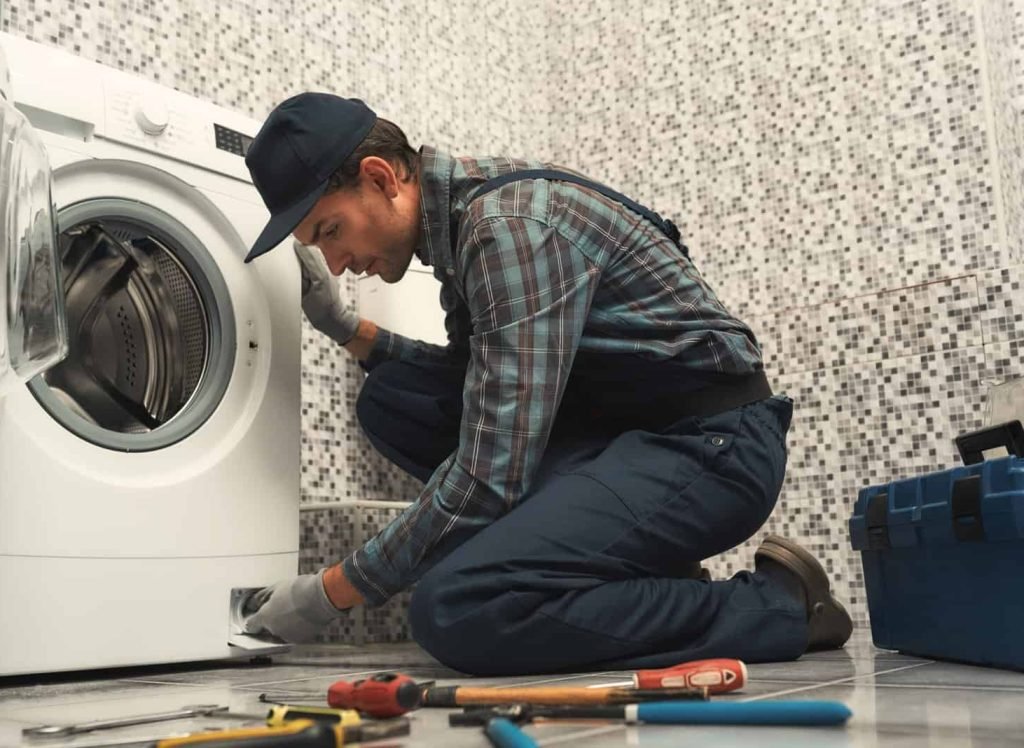 Portrait of young handyman in uniform sitting next to broken washing machine and trying to fix it. Horizontal shot. Side view