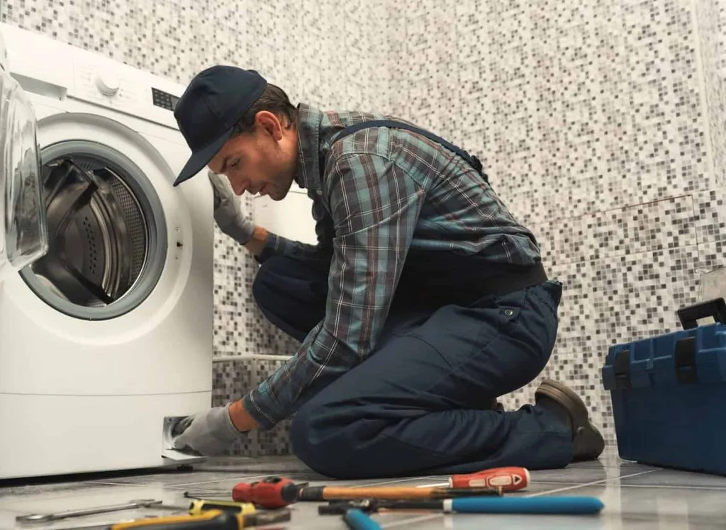 Portrait of young handyman in uniform sitting next to broken washing machine and trying to fix it. Horizontal shot. Side view