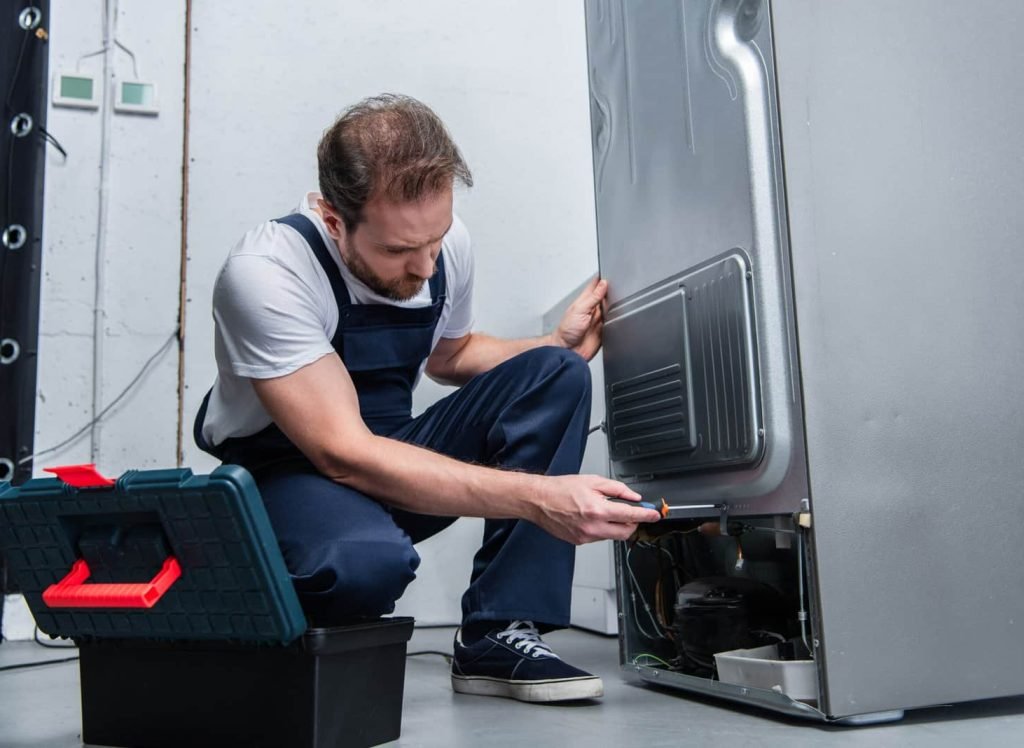 adult bearded repairman in working overall fixing refrigerator by screwdriver in kitchen